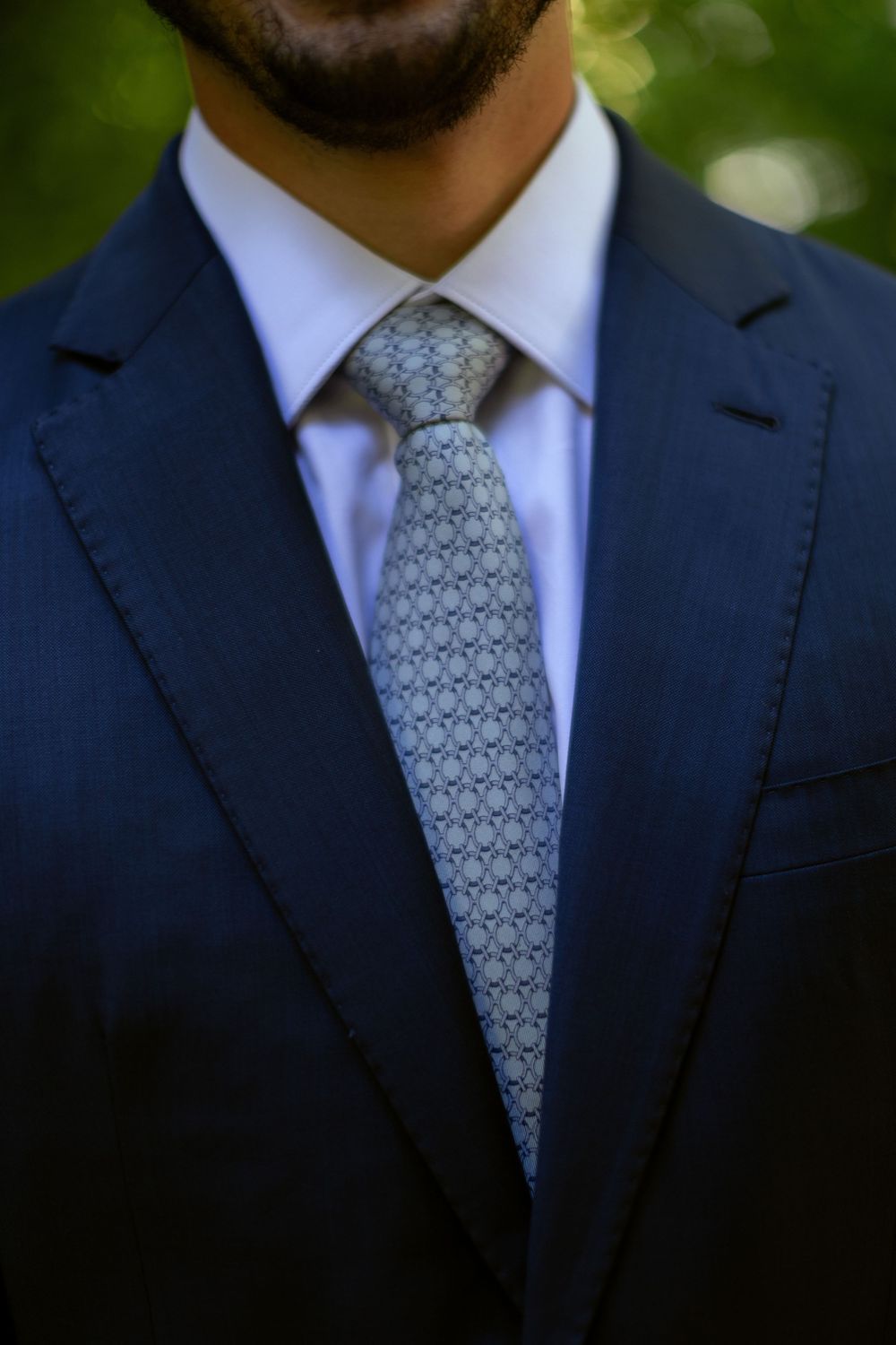 Man in navy blue suit with white dress shirt and patterned gray tie, formal business attire outdoors.