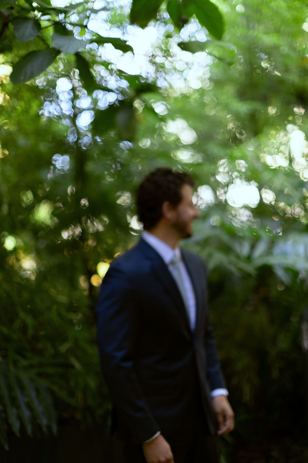 A blurred man in a dark suit and tie stands among lush green trees and foliage outdoors.