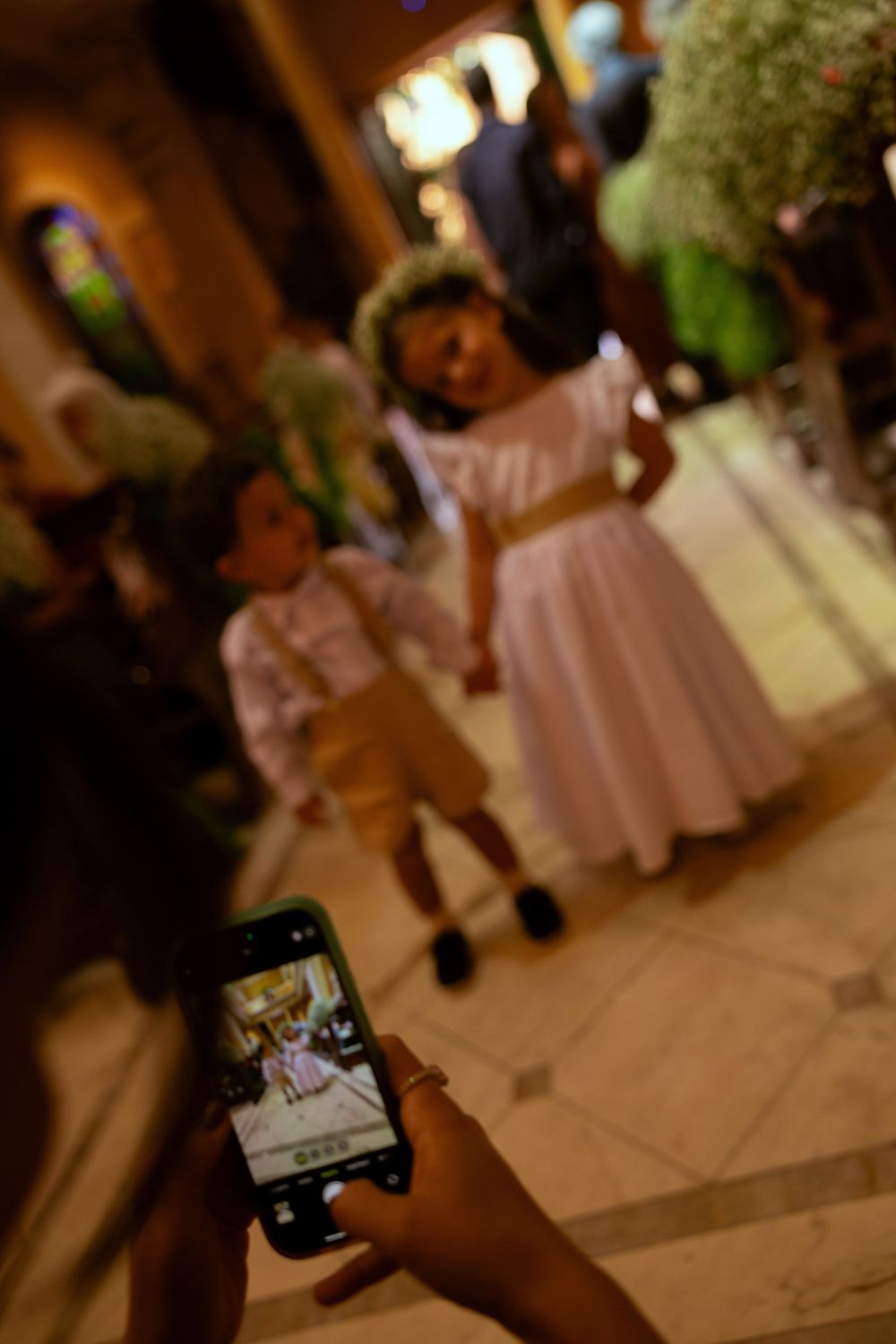 Person photographing two children in formal attire, a girl in white dress and boy in tan suit, at an elegant event.