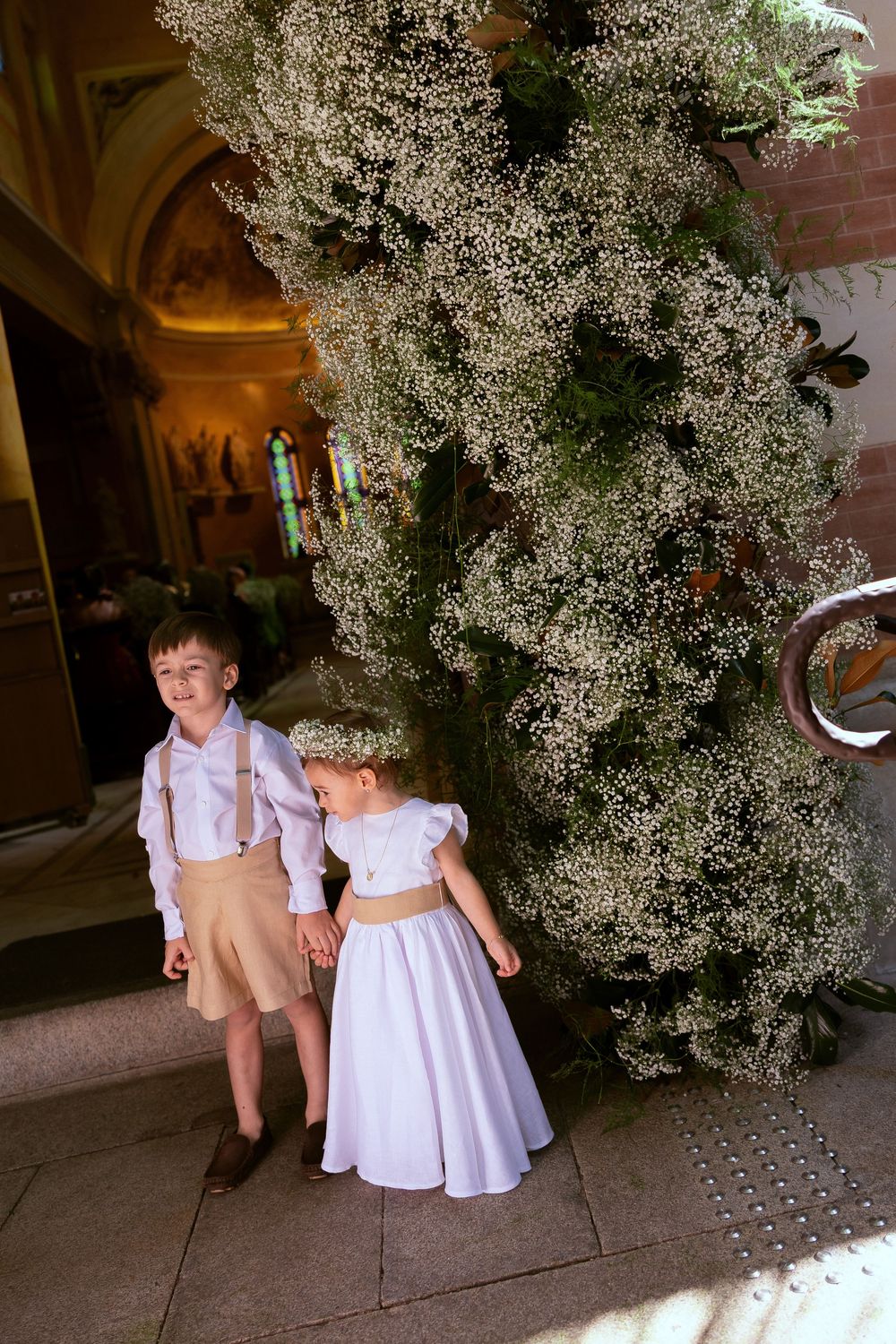 A young ring bearer and flower girl in formal attire stand beside a lush floral arch inside a church.