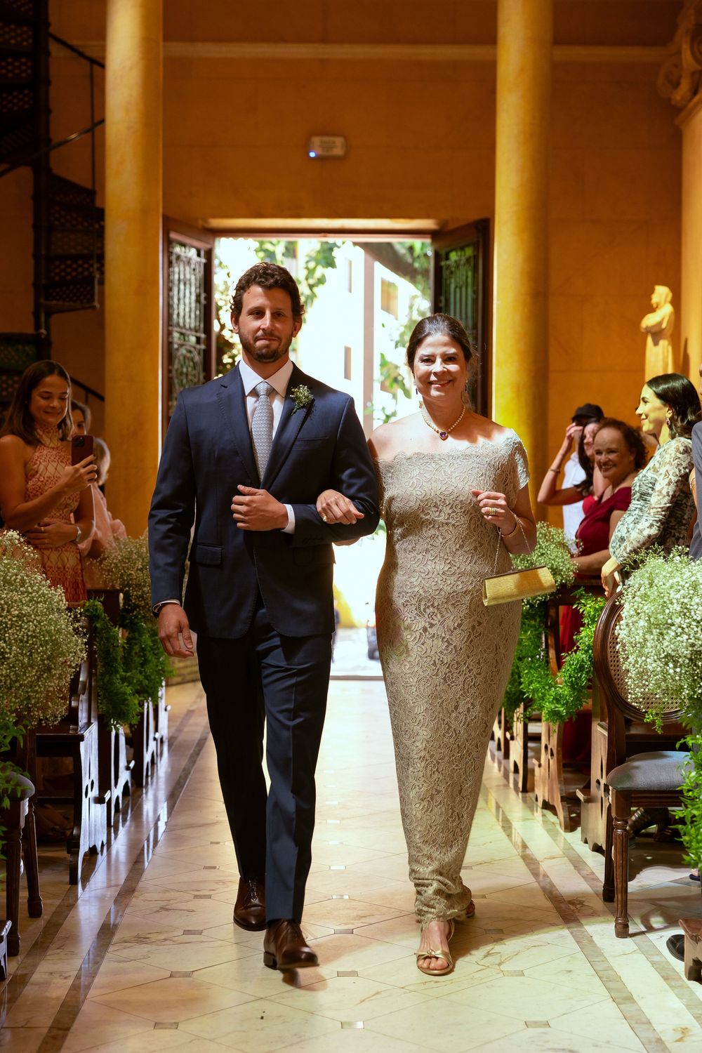 Groom in navy suit escorts elegantly dressed woman down floral-adorned wedding aisle in golden venue.