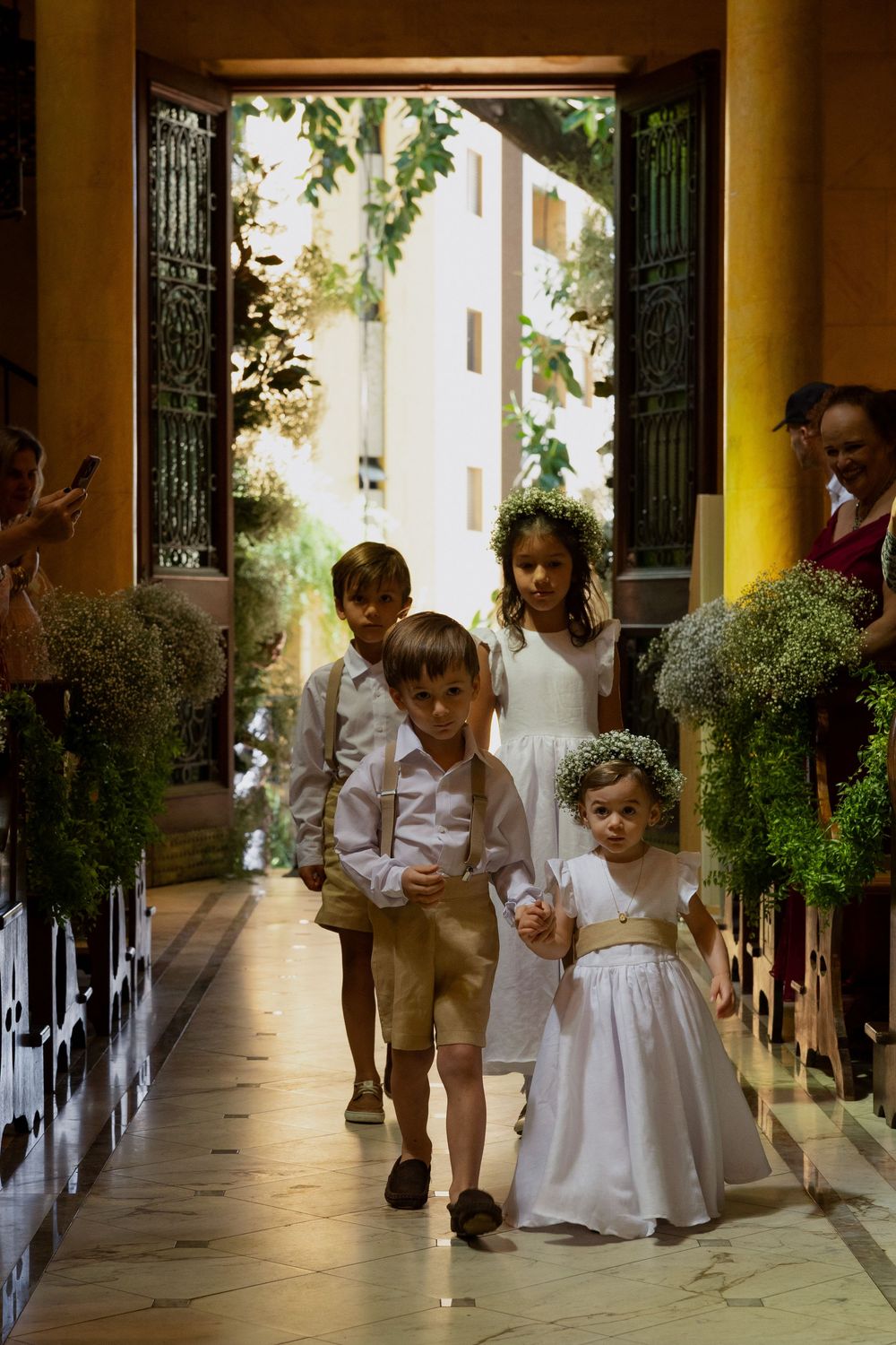 Children in formal attire walk down a church aisle as flower girls and ring bearers at a wedding ceremony.