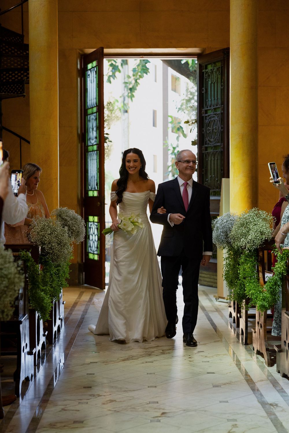 Bride in white gown walks down the aisle with her father at an elegant church wedding ceremony.