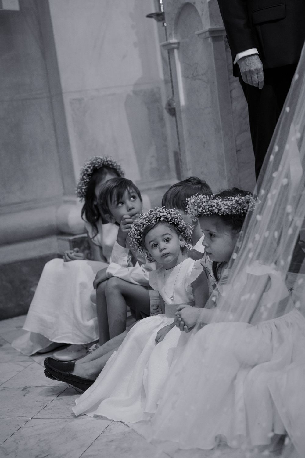 Three young flower girls in white dresses and floral crowns sit together during a wedding ceremony in black and white photo.