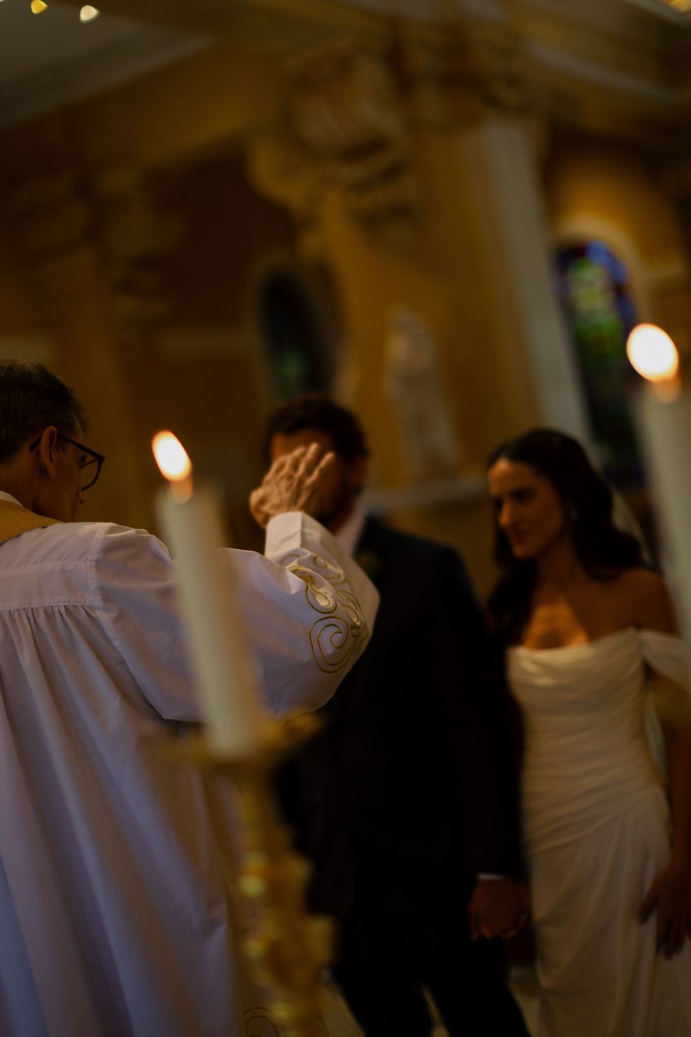 Priest blessing a bride and groom during a candlelit church wedding ceremony.