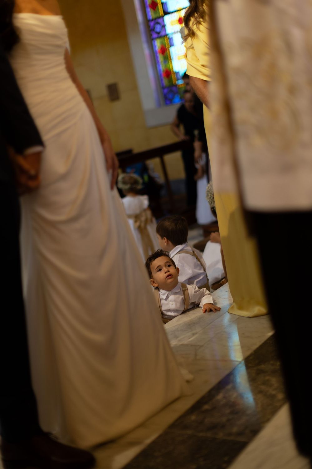Young boy in white suit lying on church floor during wedding ceremony, peeking between guests' legs.