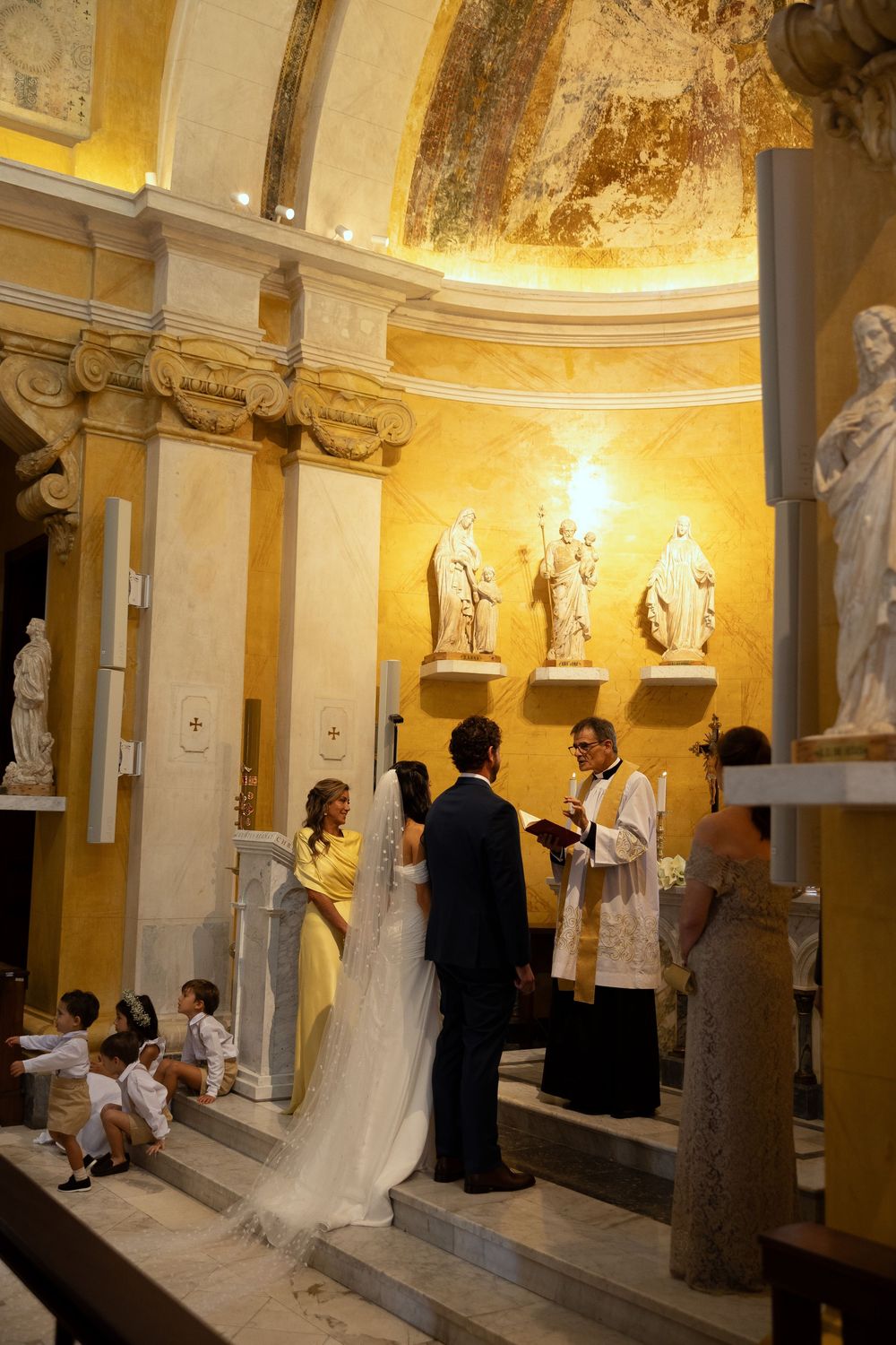 Elegant church wedding ceremony with bride in white gown, groom, and priest before ornate altar with religious statues.
