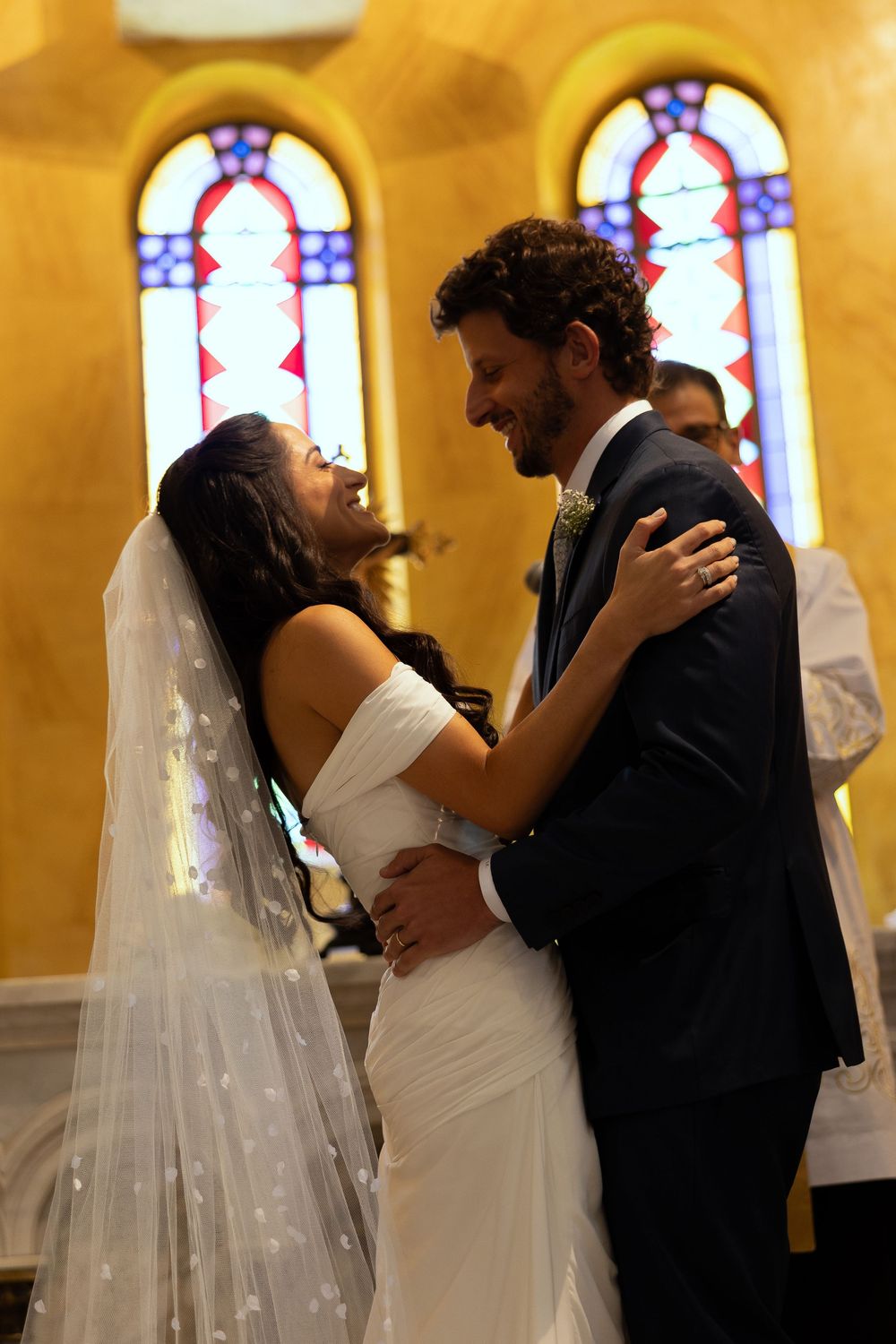 Bride and groom share a joyful embrace at church altar, stained glass windows glowing warmly behind them.