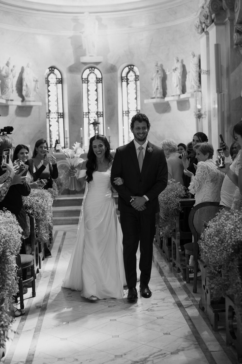 Black and white photo of a smiling bride and groom walking down the aisle of an elegant church after their wedding ceremony.