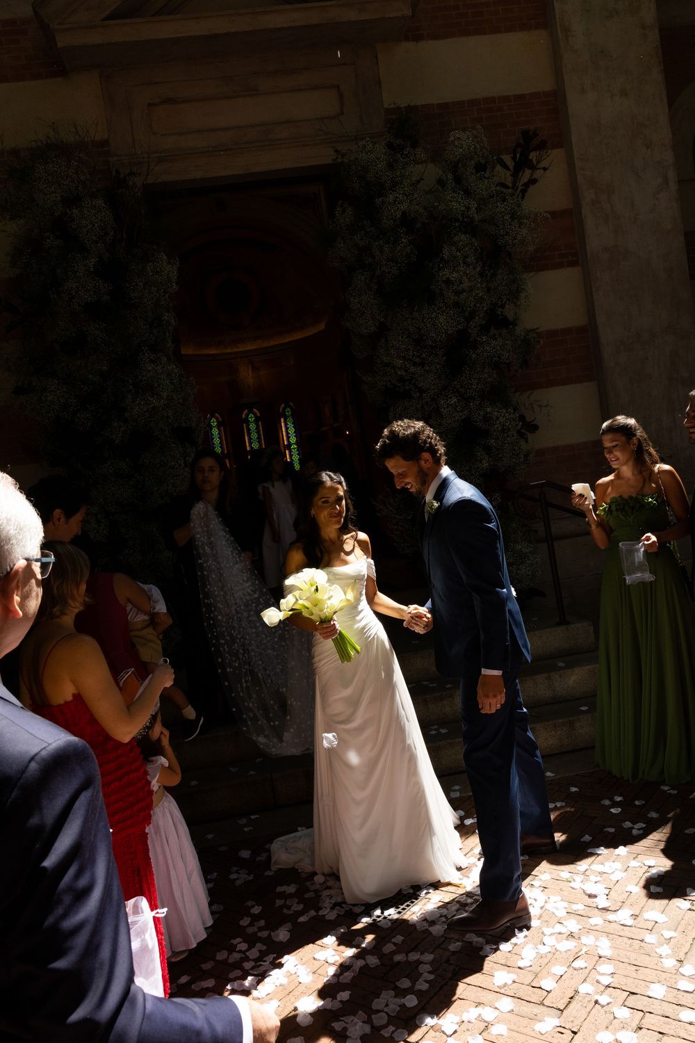Bride and groom exit church ceremony as guests celebrate with confetti on a sunny day.