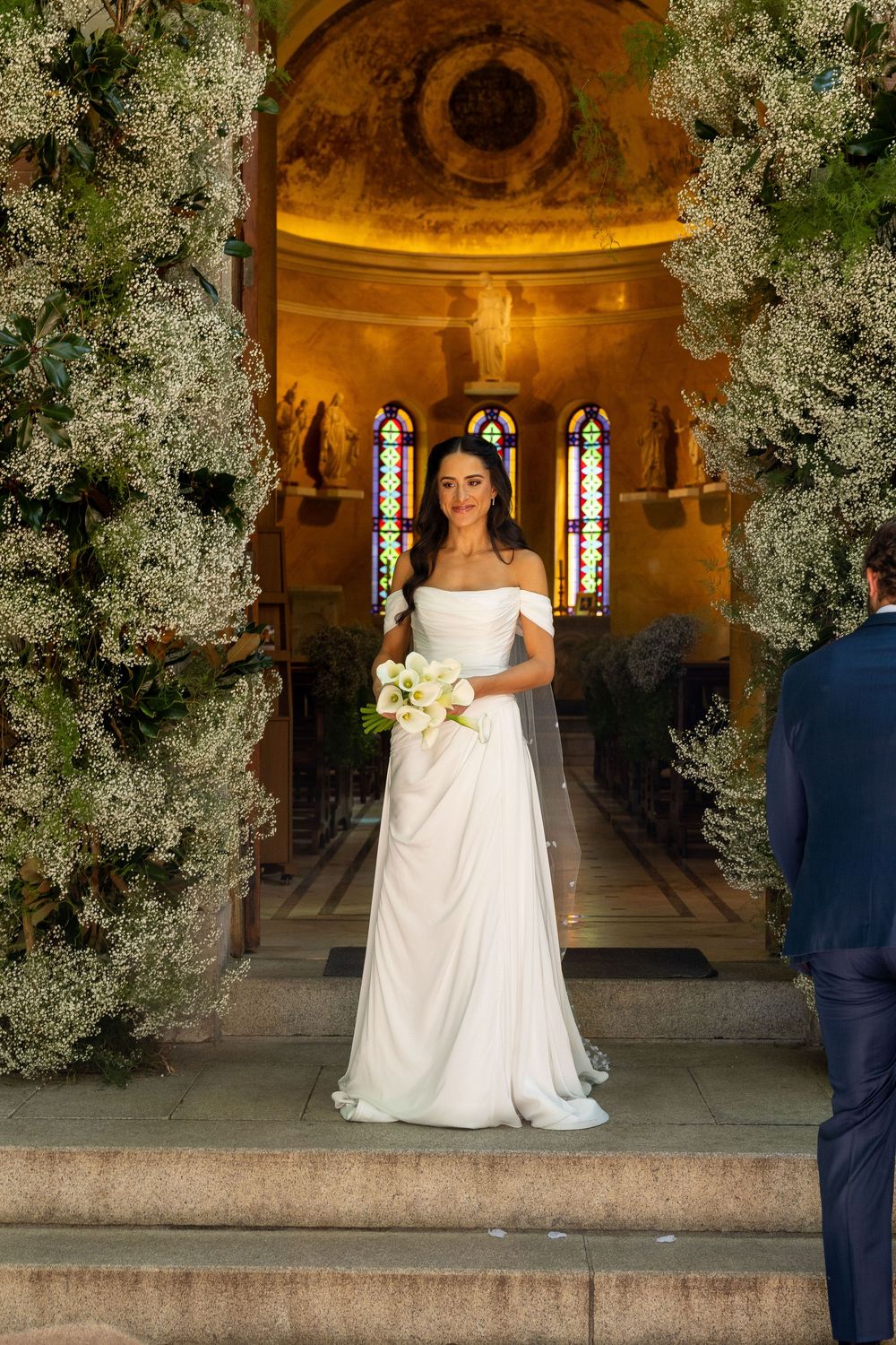 Bride in white off-shoulder gown holds bouquet at church entrance adorned with floral arrangements and stained glass windows.