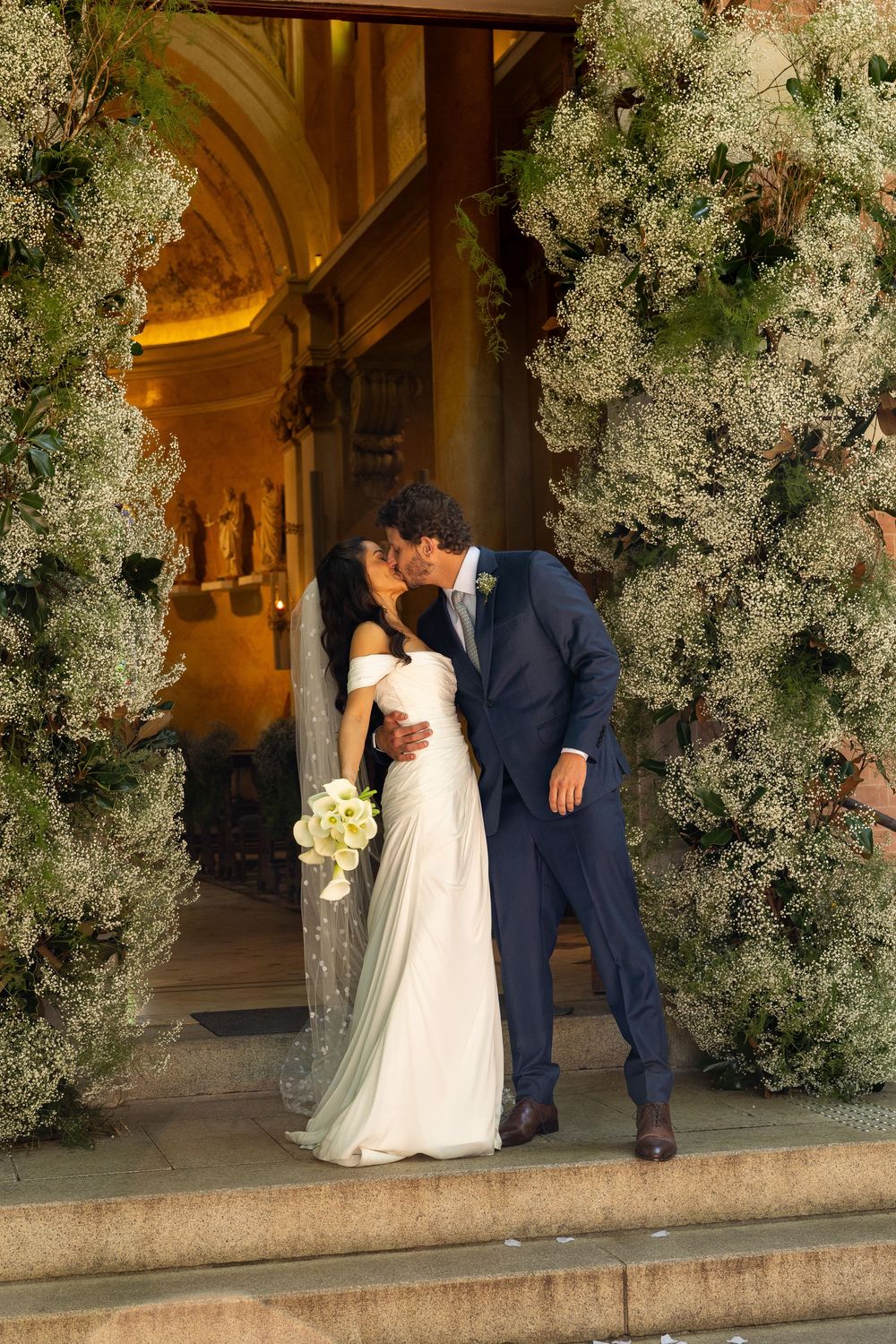 Bride and groom sharing a kiss at church entrance adorned with lush floral arrangements, bride holding white bouquet.