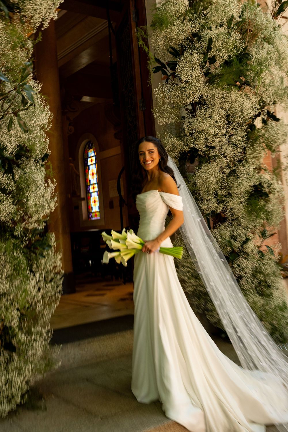 Smiling bride in off-shoulder white gown with veil holds calla lily bouquet beside lush floral arch at church entrance.