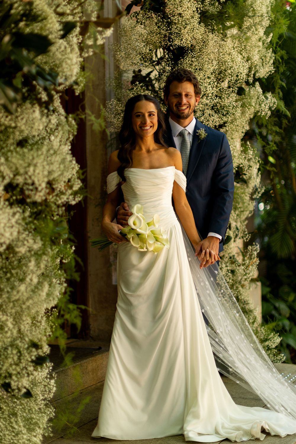 Bride in white off-shoulder gown holding calla lilies stands with groom in navy suit amid lush floral greenery.