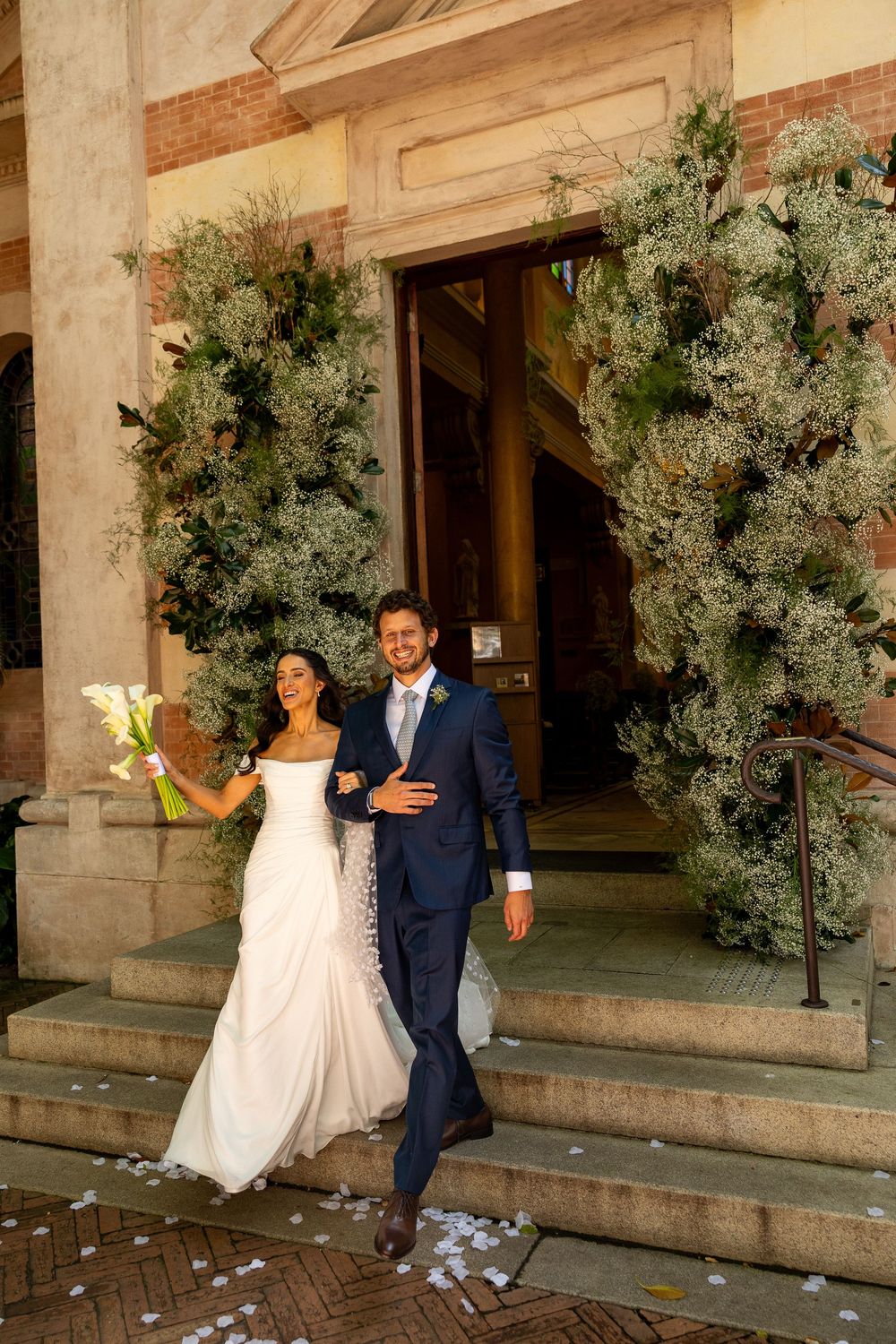 Newlywed couple exits church in wedding attire, bride holds white bouquet, groom in navy suit, floral arch backdrop.