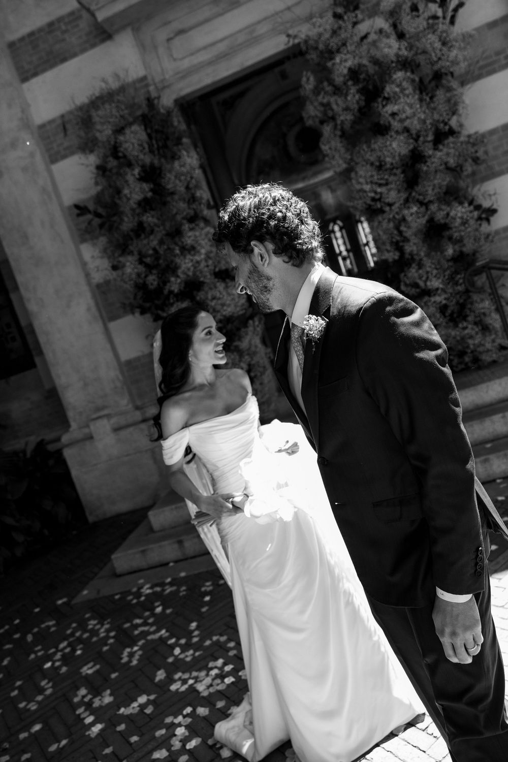 Black and white photo of a smiling bride in off-shoulder gown and groom in suit sharing a romantic moment outside a church.