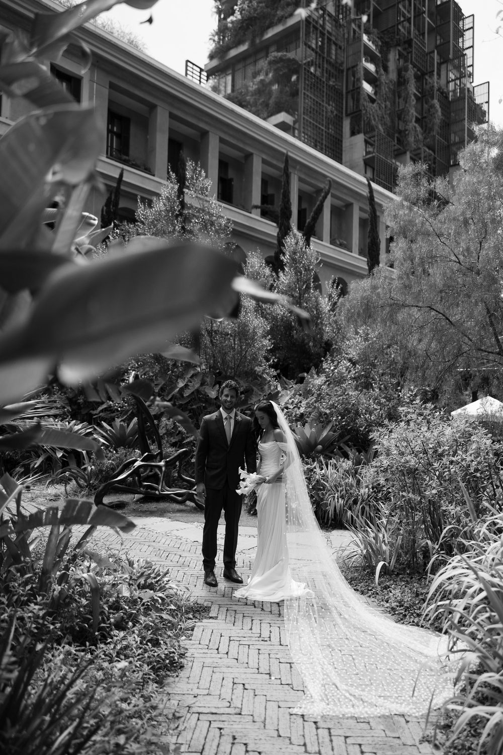 Black and white photo of a bride and groom walking on a brick path through lush gardens near an elegant building.