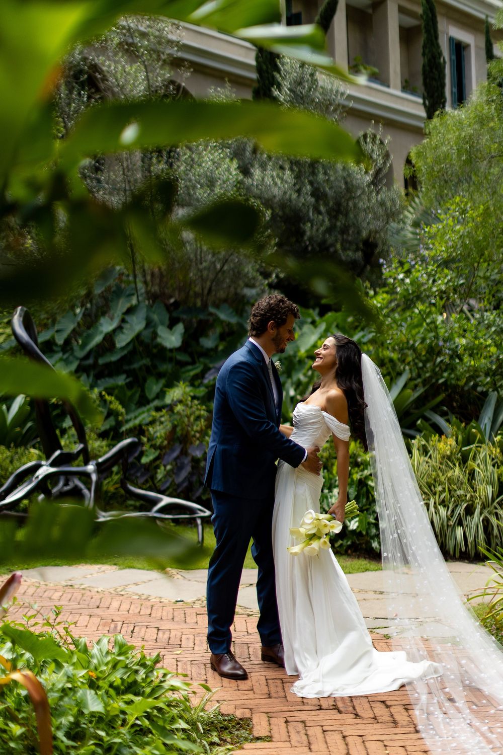 Bride and groom share a romantic moment on a brick path surrounded by lush greenery at an outdoor wedding.