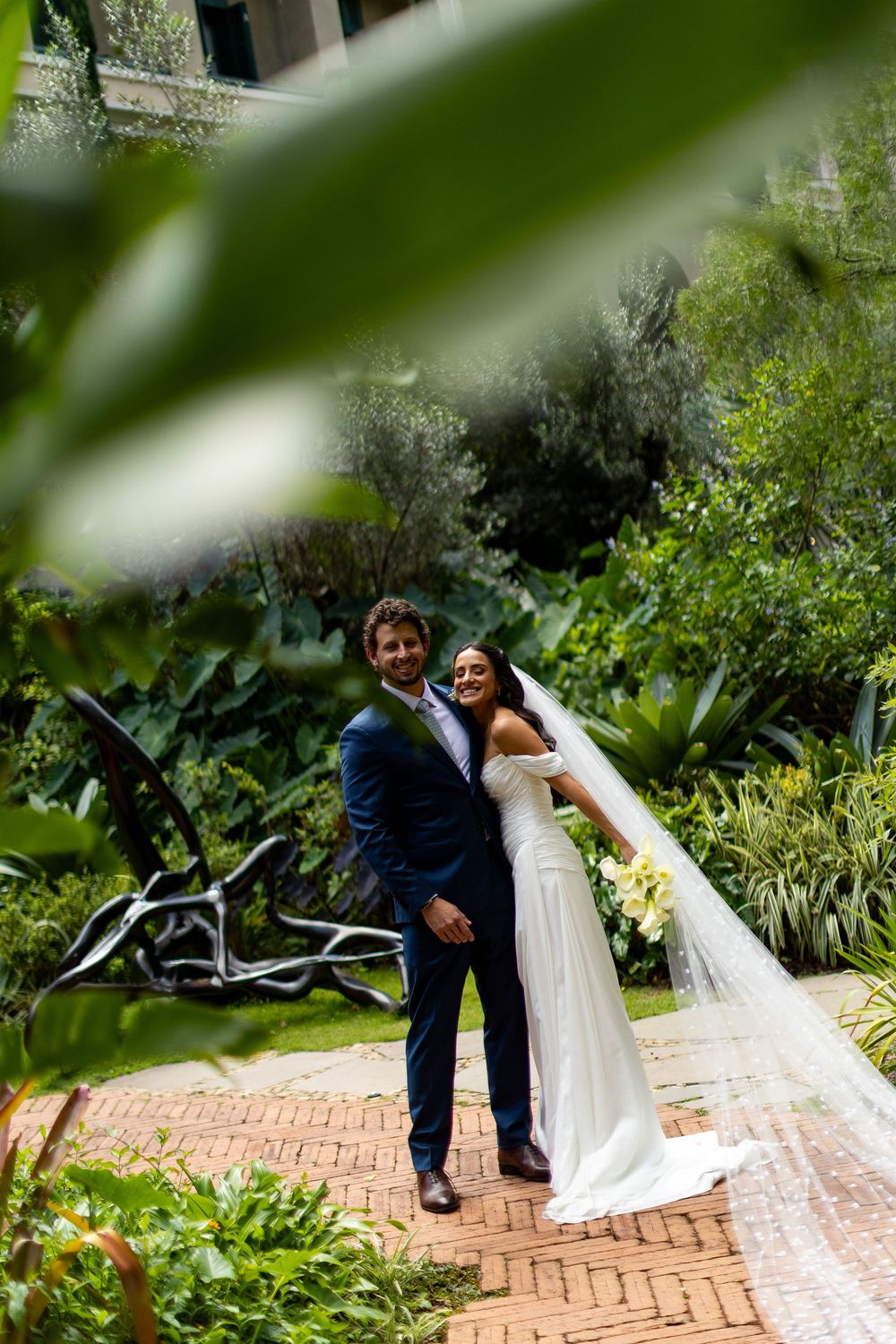 Bride and groom pose together on a brick path surrounded by lush greenery, bride wearing a white gown with veil.