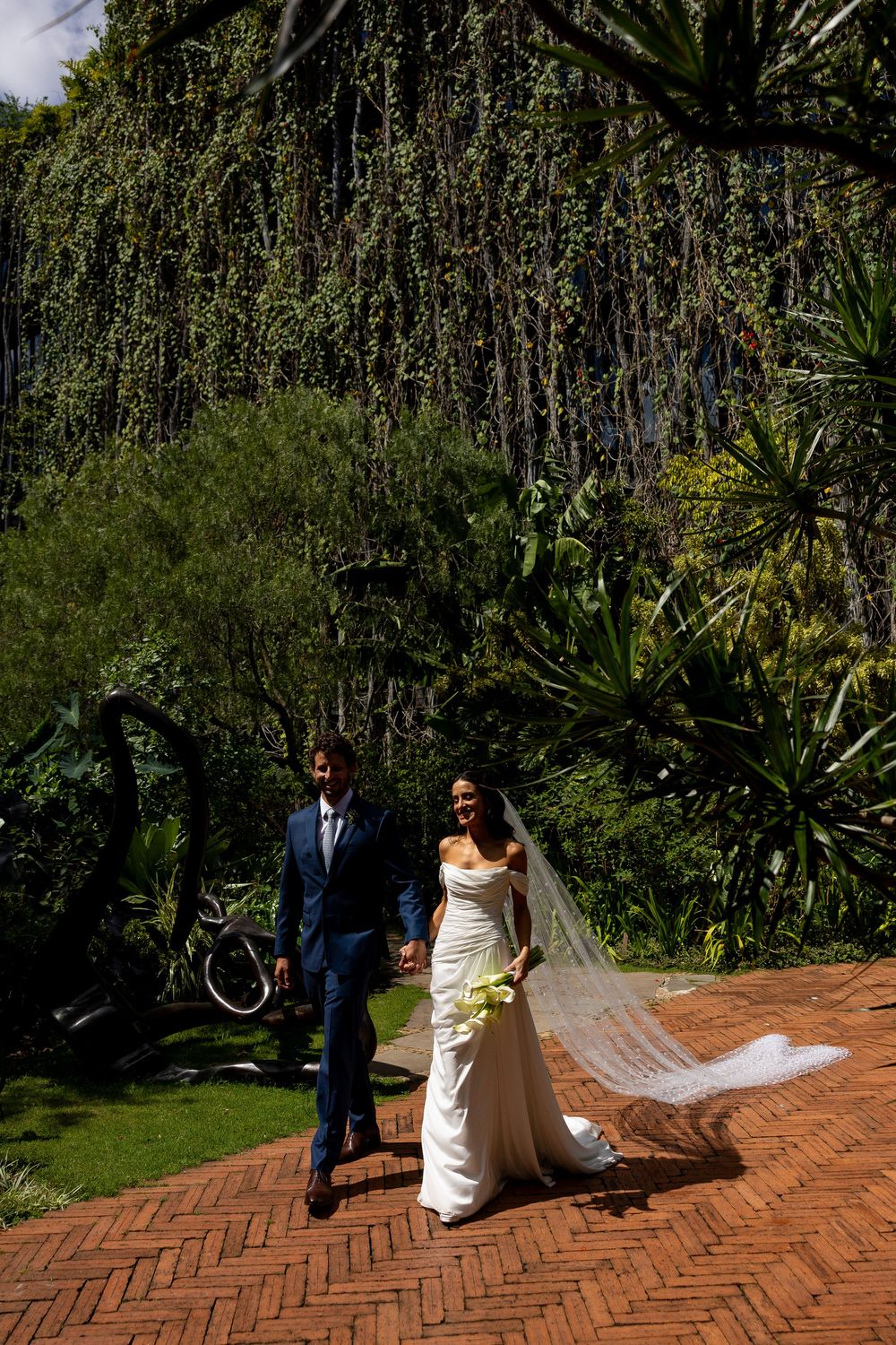 Bride in white gown with flowing veil walks hand-in-hand with groom in blue suit on brick path surrounded by lush greenery.