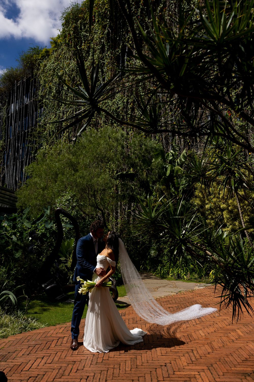 Bride and groom share a romantic kiss outdoors, with veil flowing in wind, surrounded by lush tropical greenery.