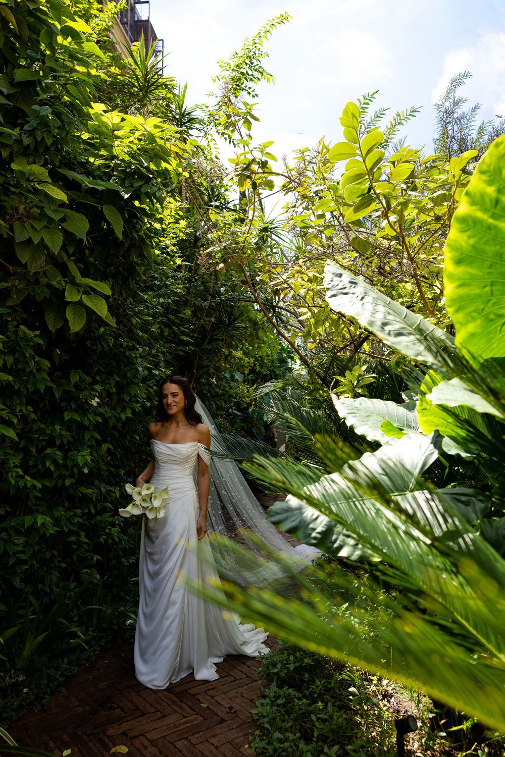 Bride in white gown and veil holding bouquet stands in lush green garden pathway on her wedding day.