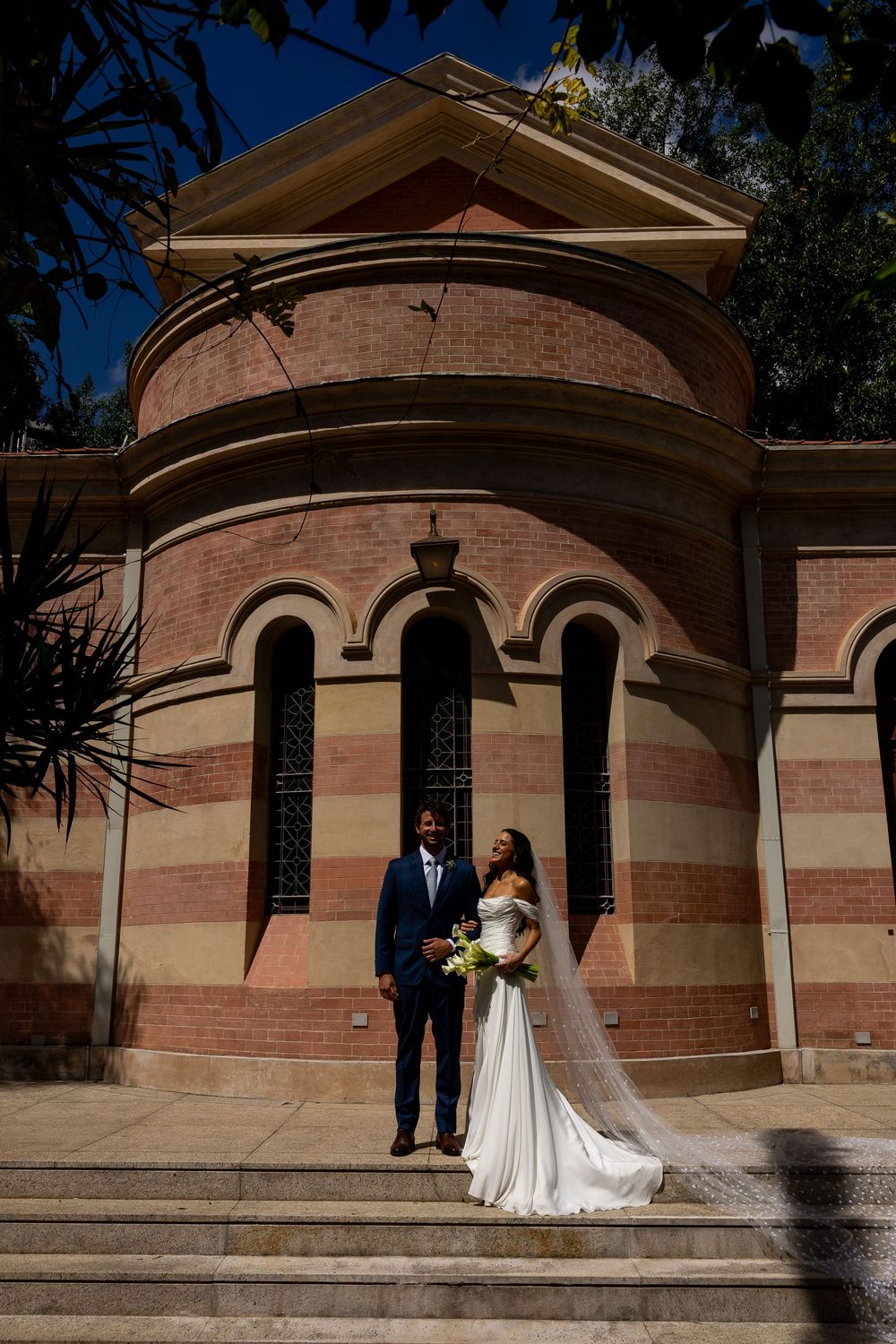 Bride and groom posing on steps before a historic red brick church with arched windows on their wedding day.