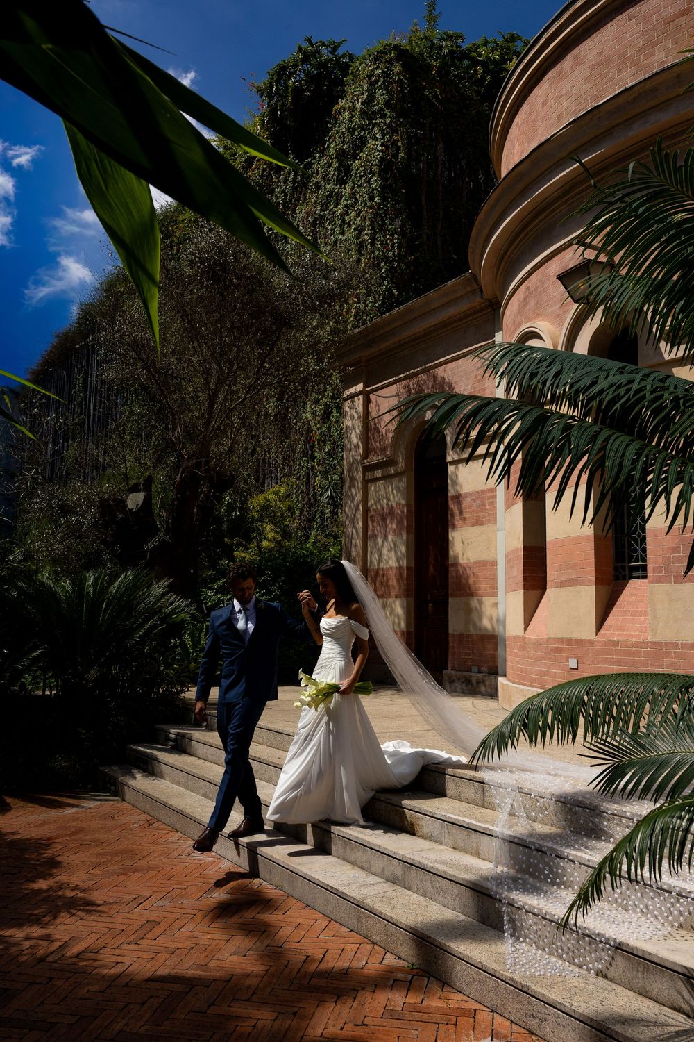 Bride and groom walking up brick steps beside a lush ivy-covered brick building surrounded by tropical palms on a sunny day.