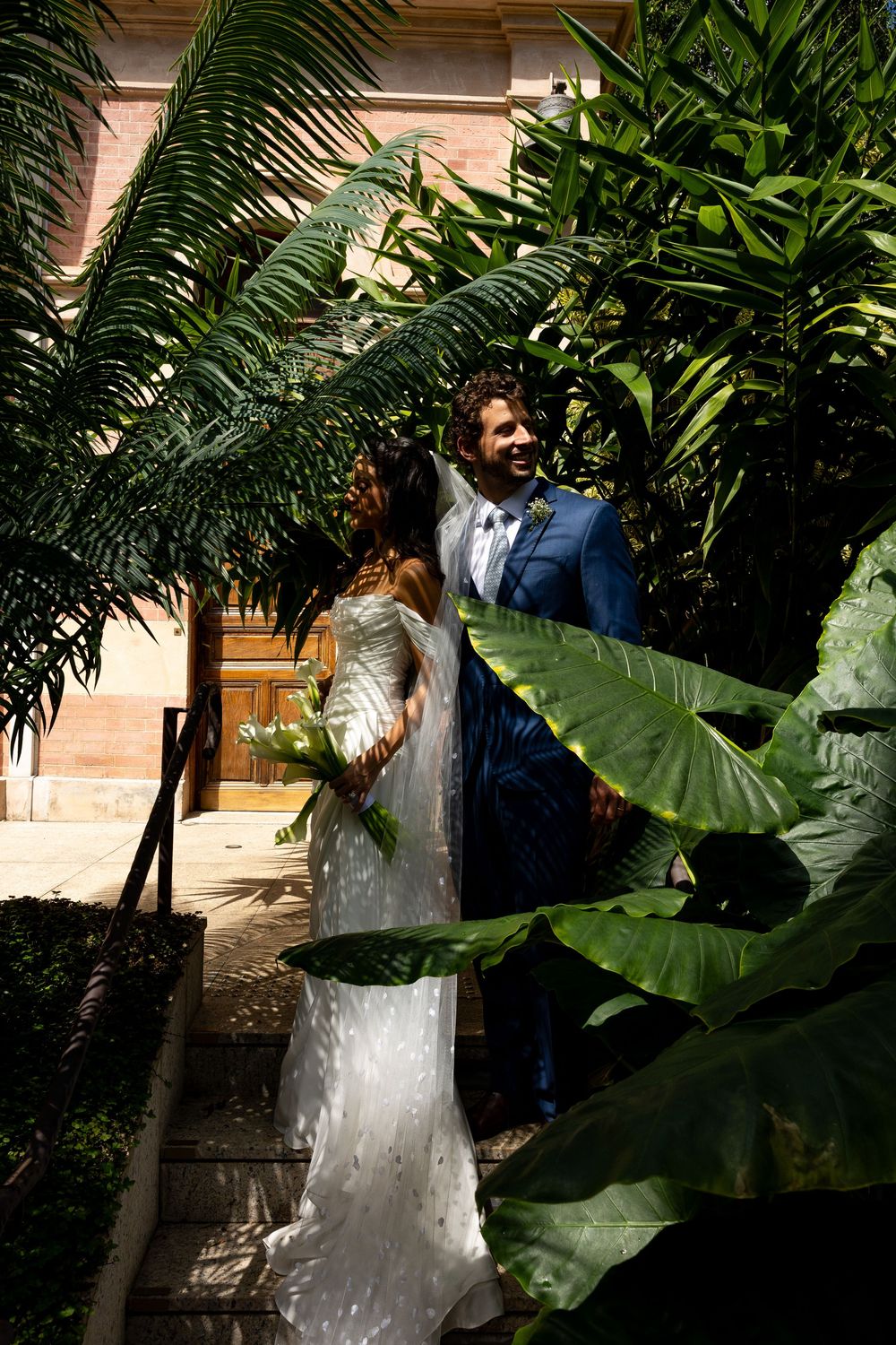 Bride in white gown and groom in blue suit stand among lush tropical plants outside a building.