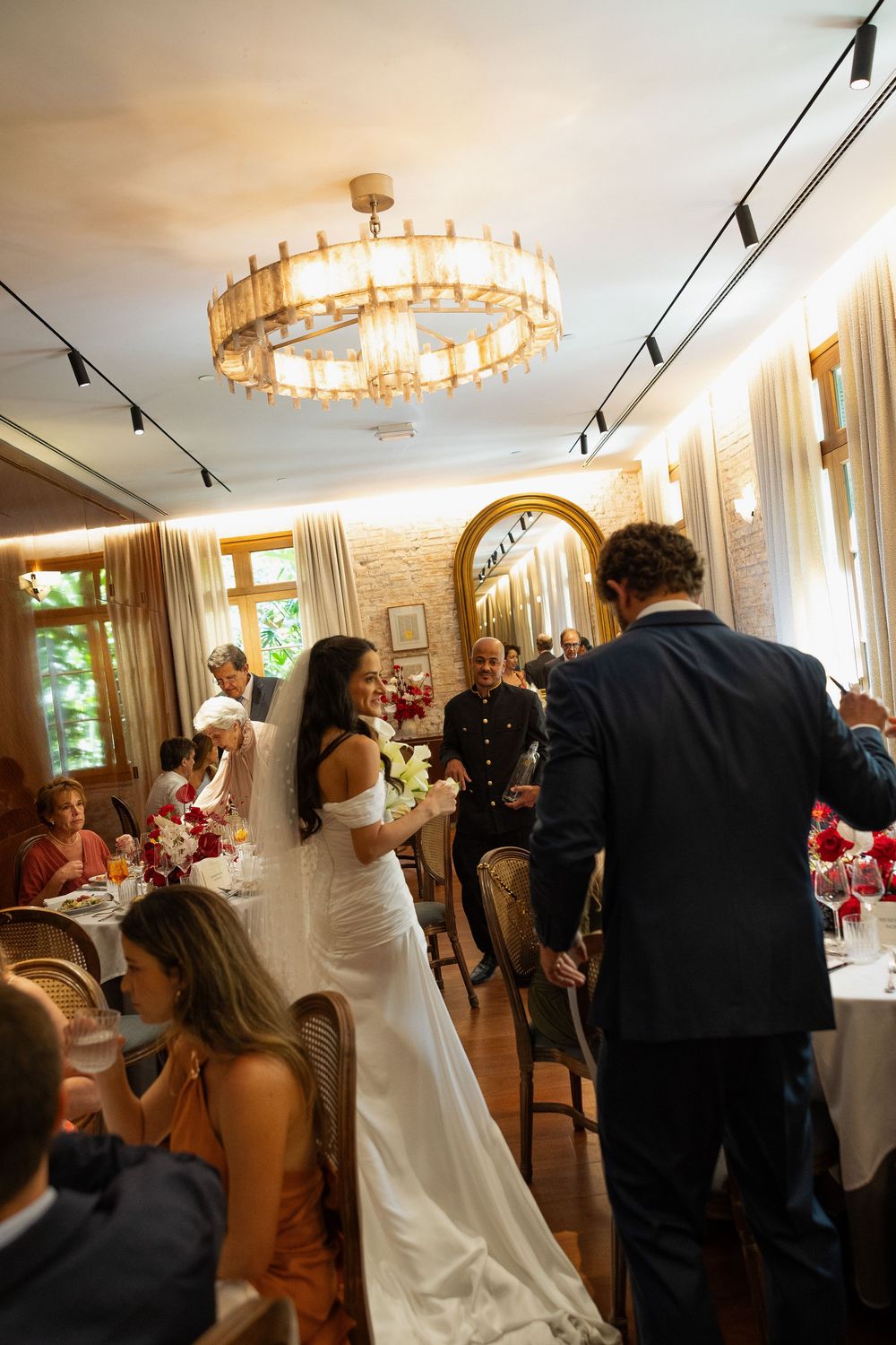 Bride and groom walk through elegant wedding reception hall with chandelier and seated guests.