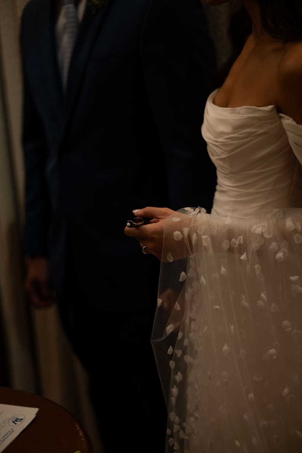 Bride in white off-shoulder floral tulle wedding gown holding phone beside groom in dark suit.