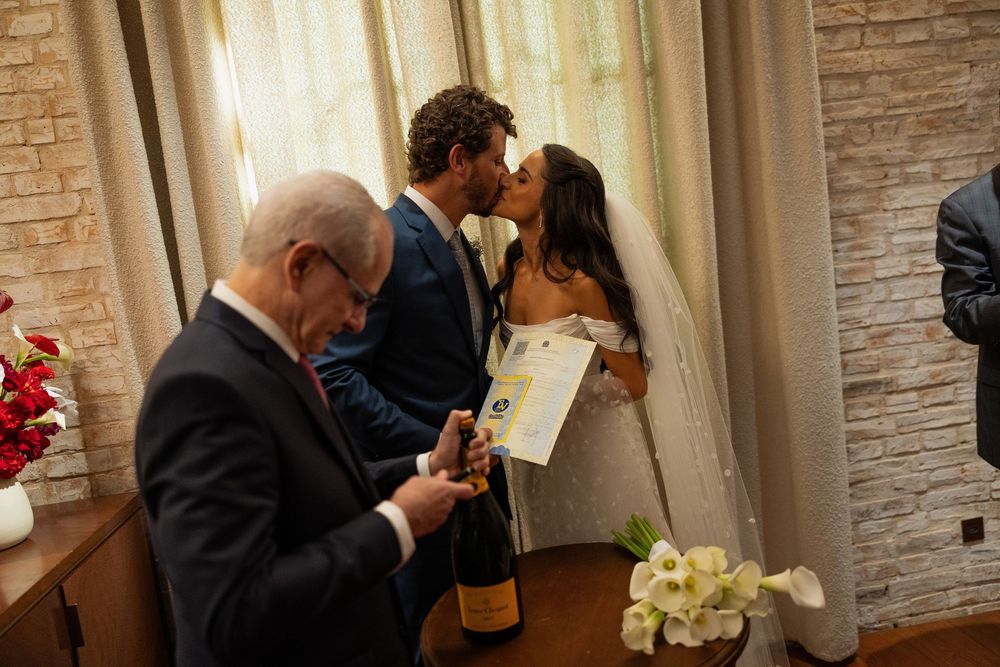 Bride and groom share a kiss during intimate wedding ceremony while officiant holds champagne bottle and documents.