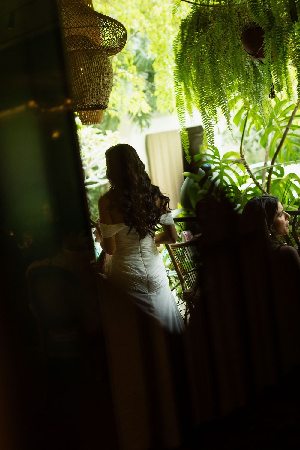 Bride in white off-shoulder gown stands among lush tropical greenery and hanging rattan lanterns in elegant venue.