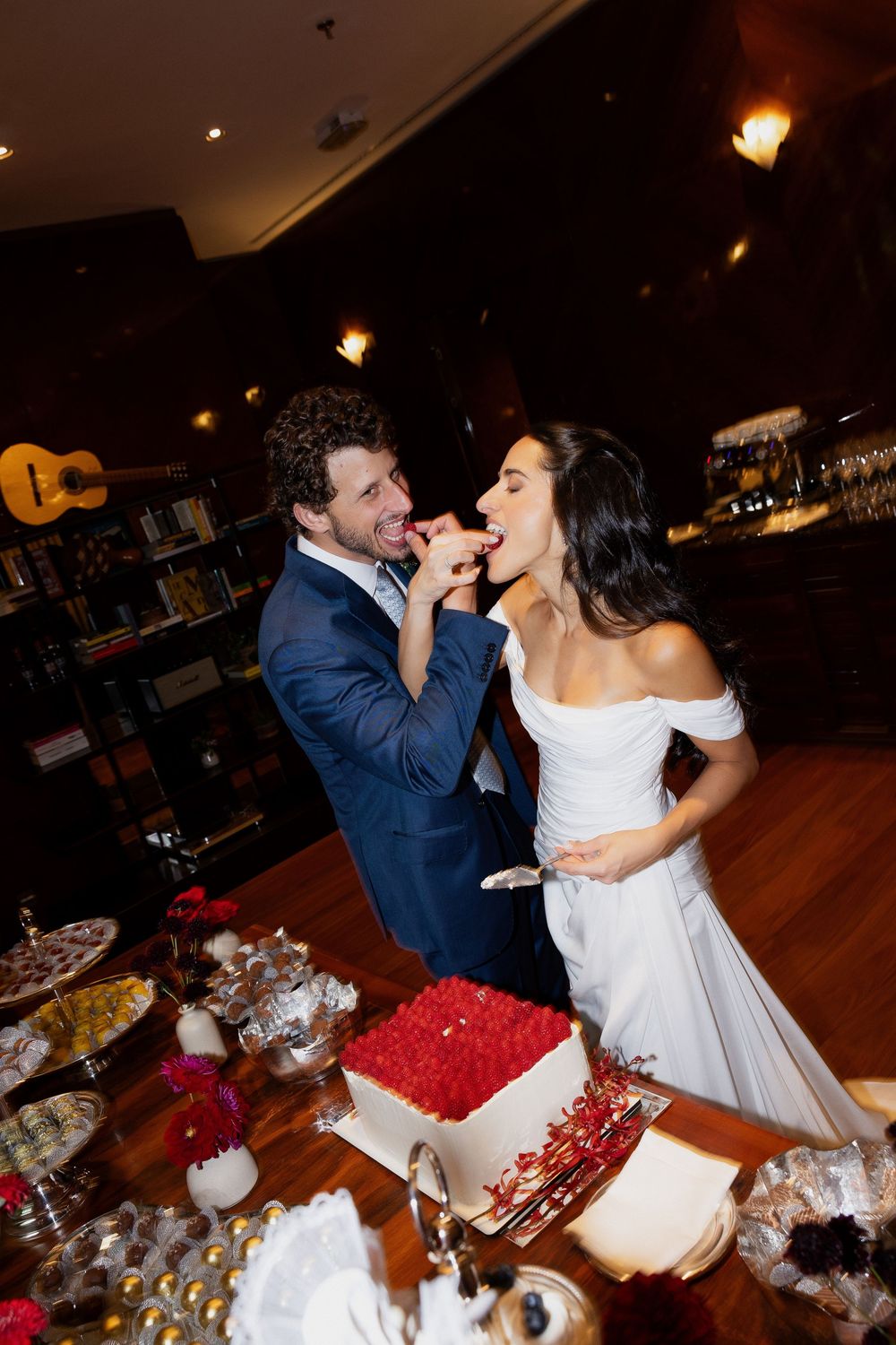 Bride and groom feeding each other wedding cake at reception table with white rose-decorated cake.