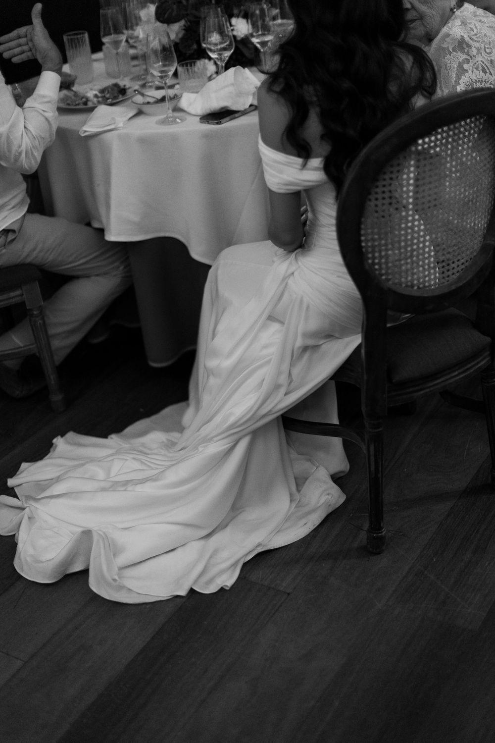Black and white photo of a bride in an elegant off-shoulder gown seated at a wedding reception table.