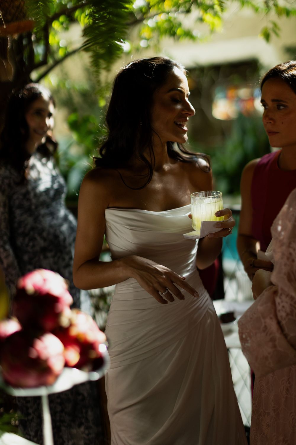 Bride in white strapless gown holding a yellow drink at an outdoor wedding reception with guests.