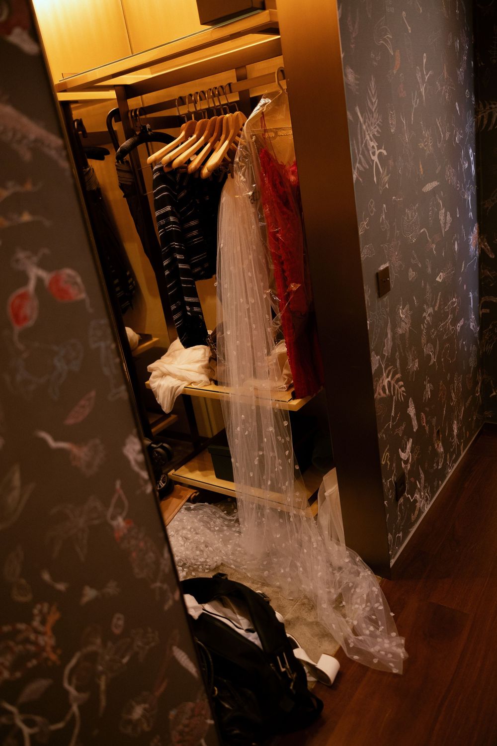 Open wardrobe closet with hanging clothes, a white bridal veil, and shoes on the floor in a dimly lit room.