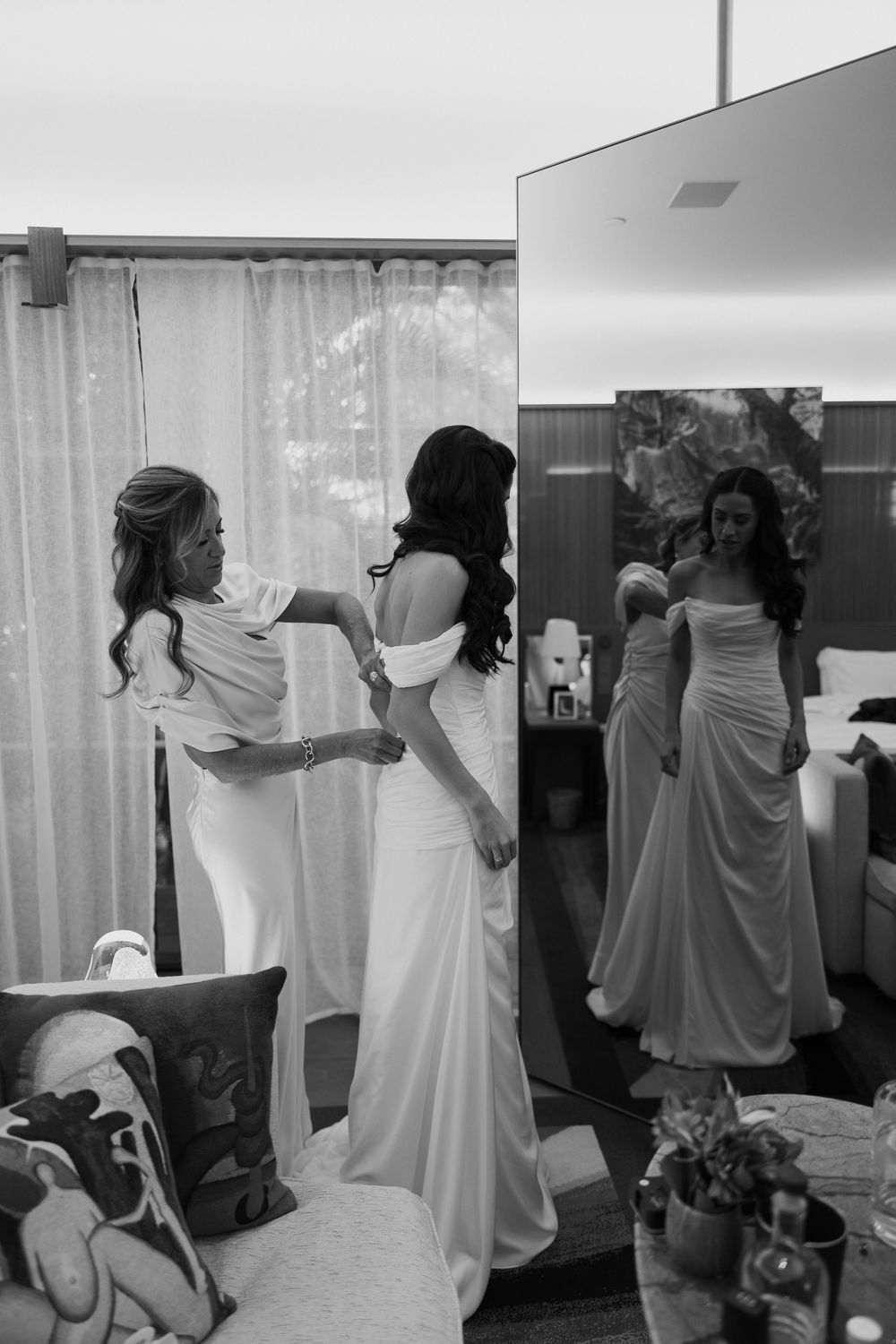 Bride being helped into white off-shoulder wedding gown by bridesmaid, reflected in large mirror, black and white photo.