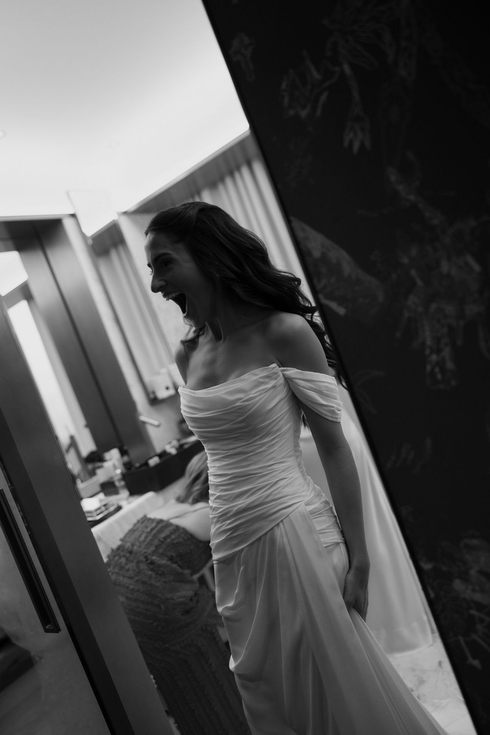 Black and white photo of a laughing bride in an off-shoulder wedding gown, captured in a candid moment indoors.