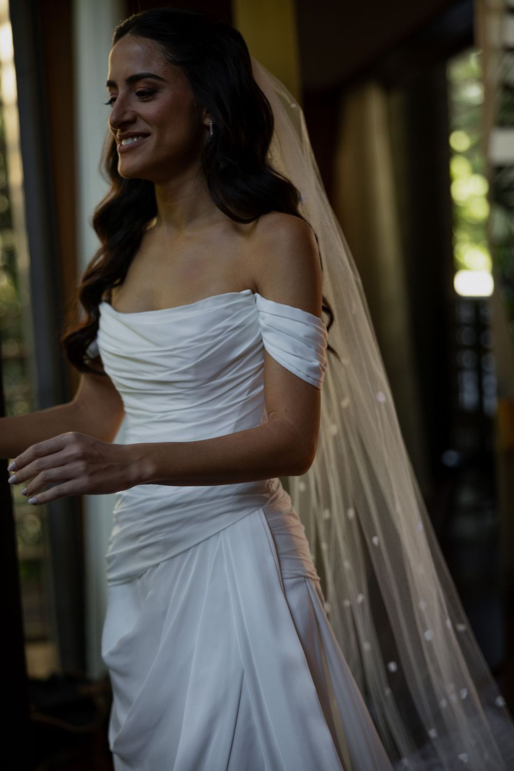 Smiling bride in off-shoulder white satin gown with pearl-dotted veil, dark wavy hair, standing in soft natural light.