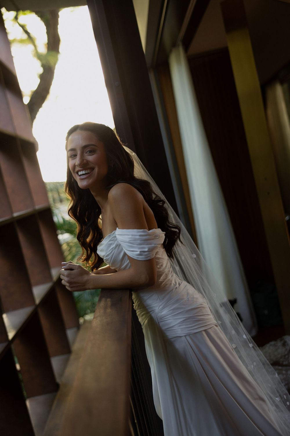 Smiling bride in white off-shoulder gown and veil leans on railing in golden sunlight on outdoor balcony.