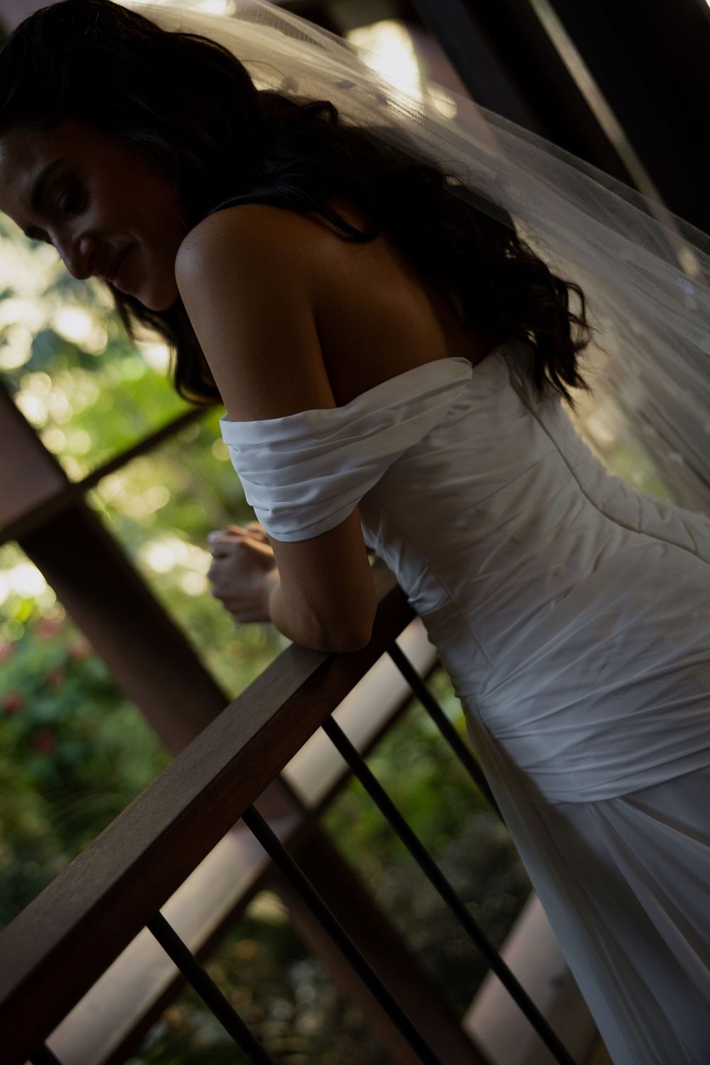 Bride in off-shoulder white wedding dress and veil leaning on balcony railing surrounded by lush greenery.