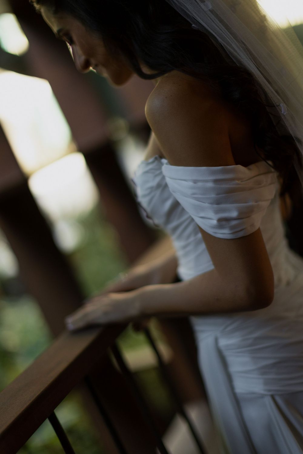 Bride in white off-shoulder gown and veil leans gracefully on a wooden railing outdoors.