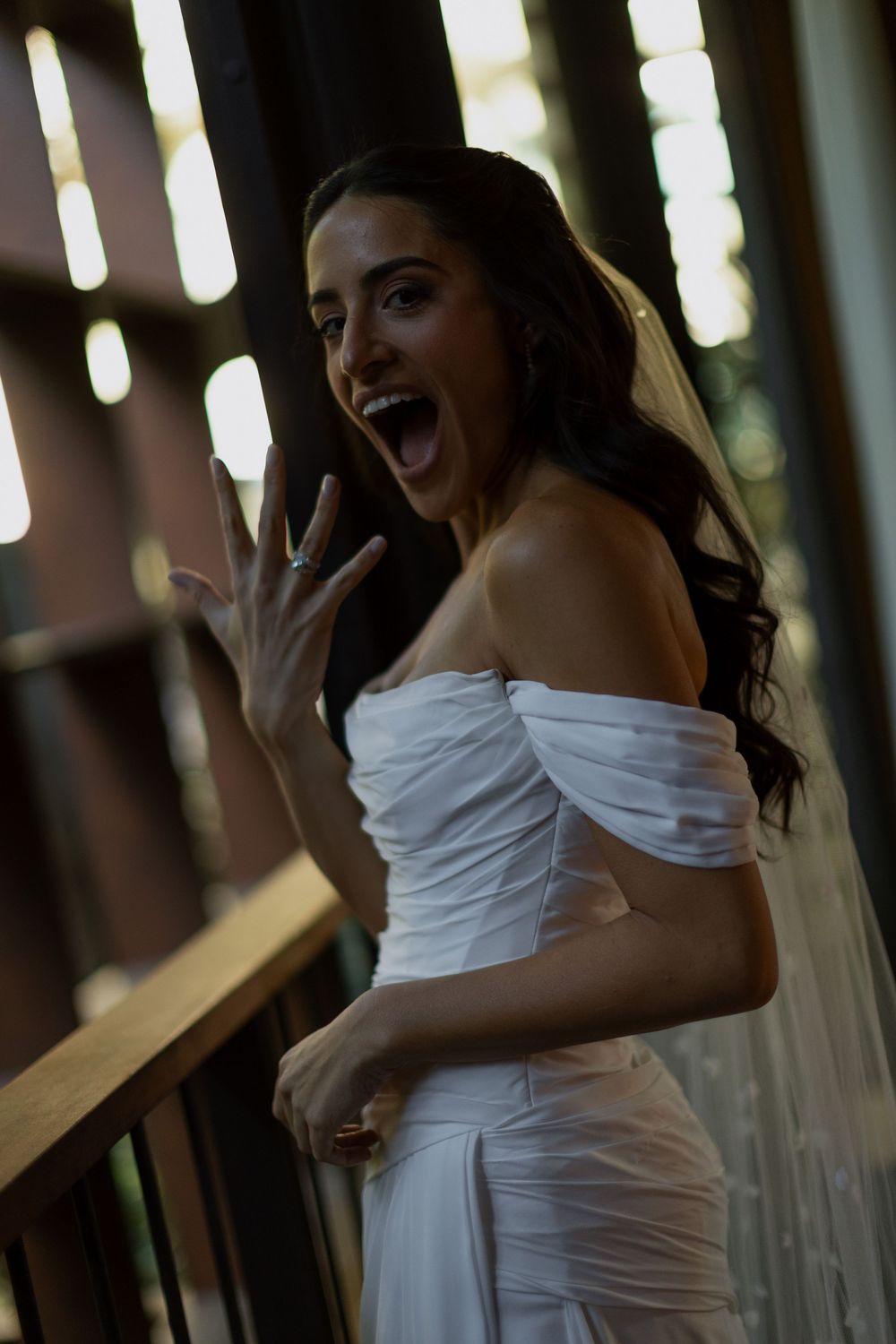 Excited bride in white off-shoulder gown and veil, showing ring with open-mouthed expression on balcony.