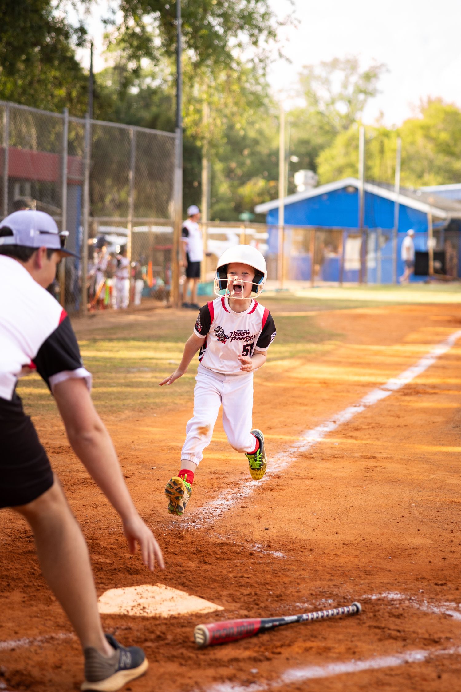 Young baseball player in white uniform runs bases with excitement during a youth league game on a red dirt field.
