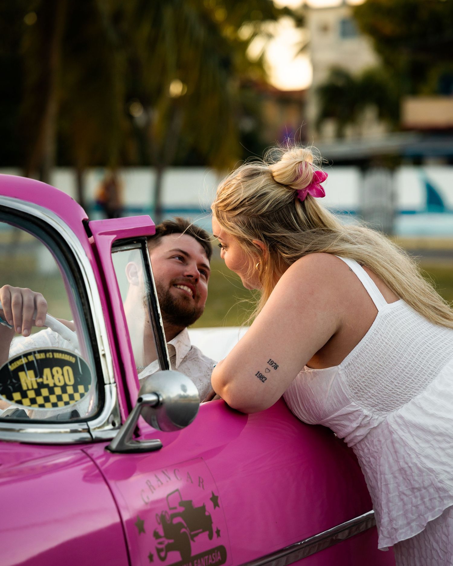 Couple sharing a romantic moment beside a vintage pink taxi car at sunset.