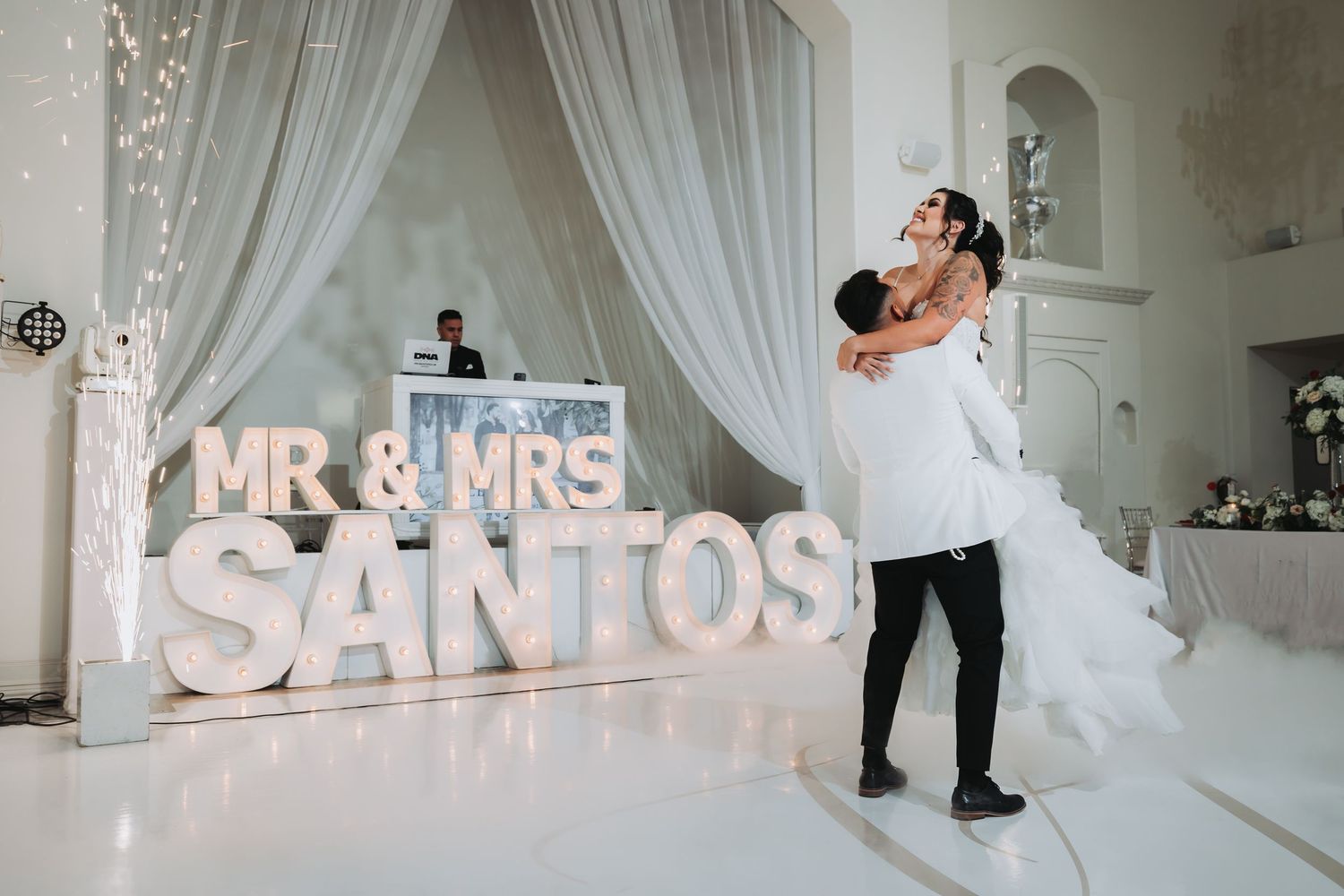 Bride and groom on a dance floor, he lifts her up by a 'Mr. & Mrs.' lit letter sign. Cold sparks fly as a DJ works in the background.