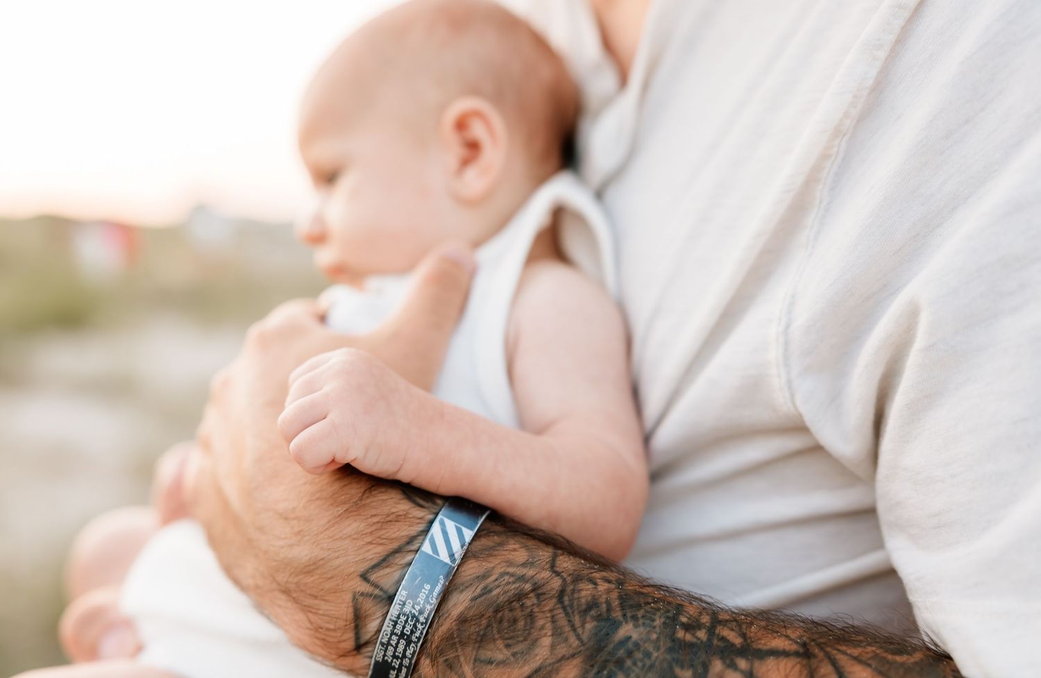 Tattooed father tenderly holding a newborn baby against his chest outdoors in soft natural light.