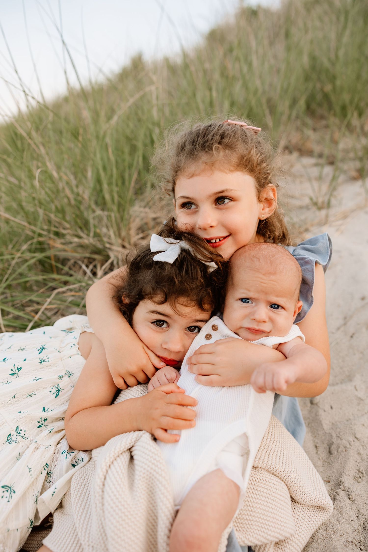 Three young siblings cuddle together on a sandy beach, with the oldest girl smiling and hugging her younger sister and baby brother.