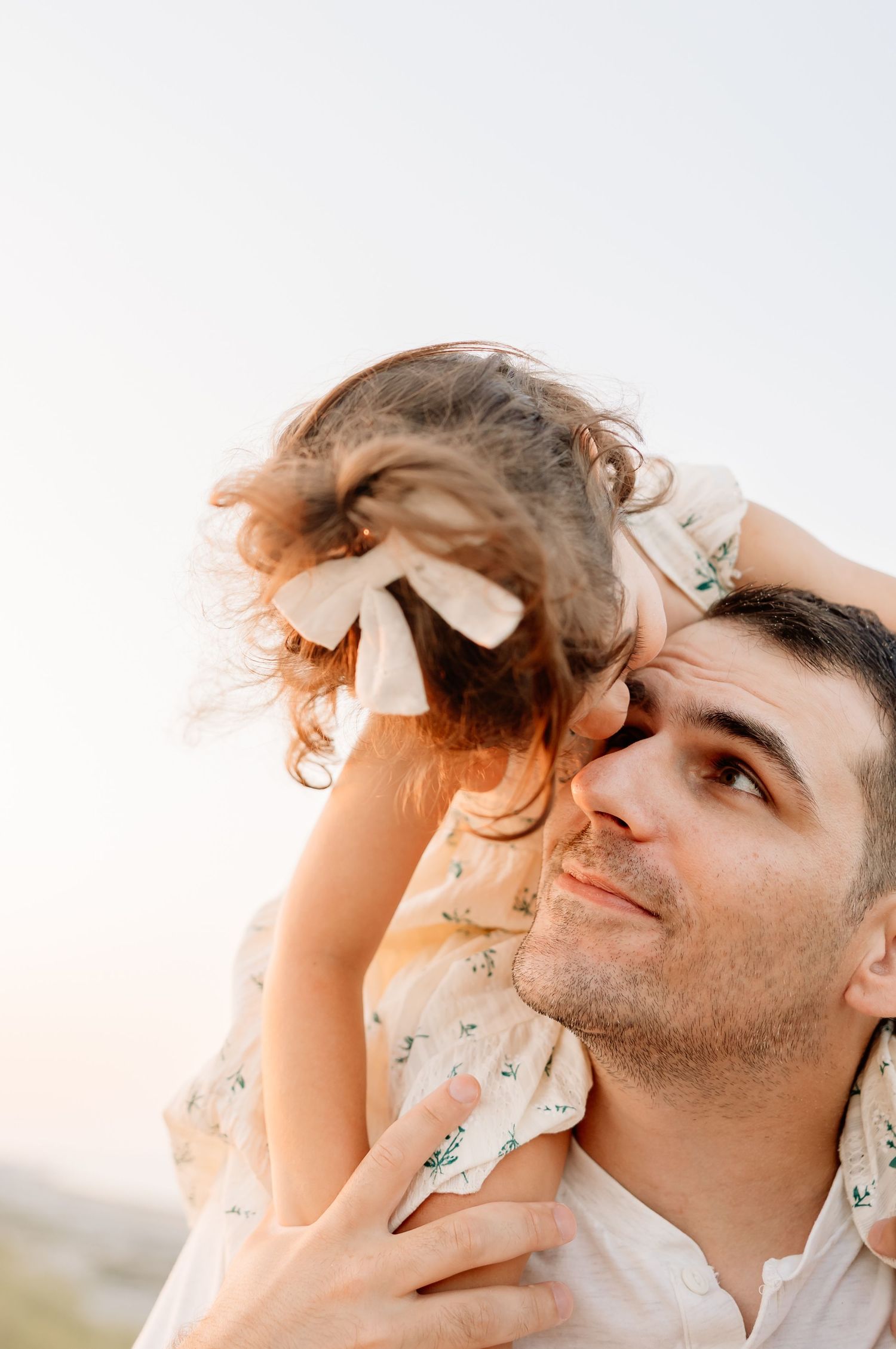 A father lovingly holds his young daughter on his shoulders outdoors, smiling up at her as her hair blows in the wind.